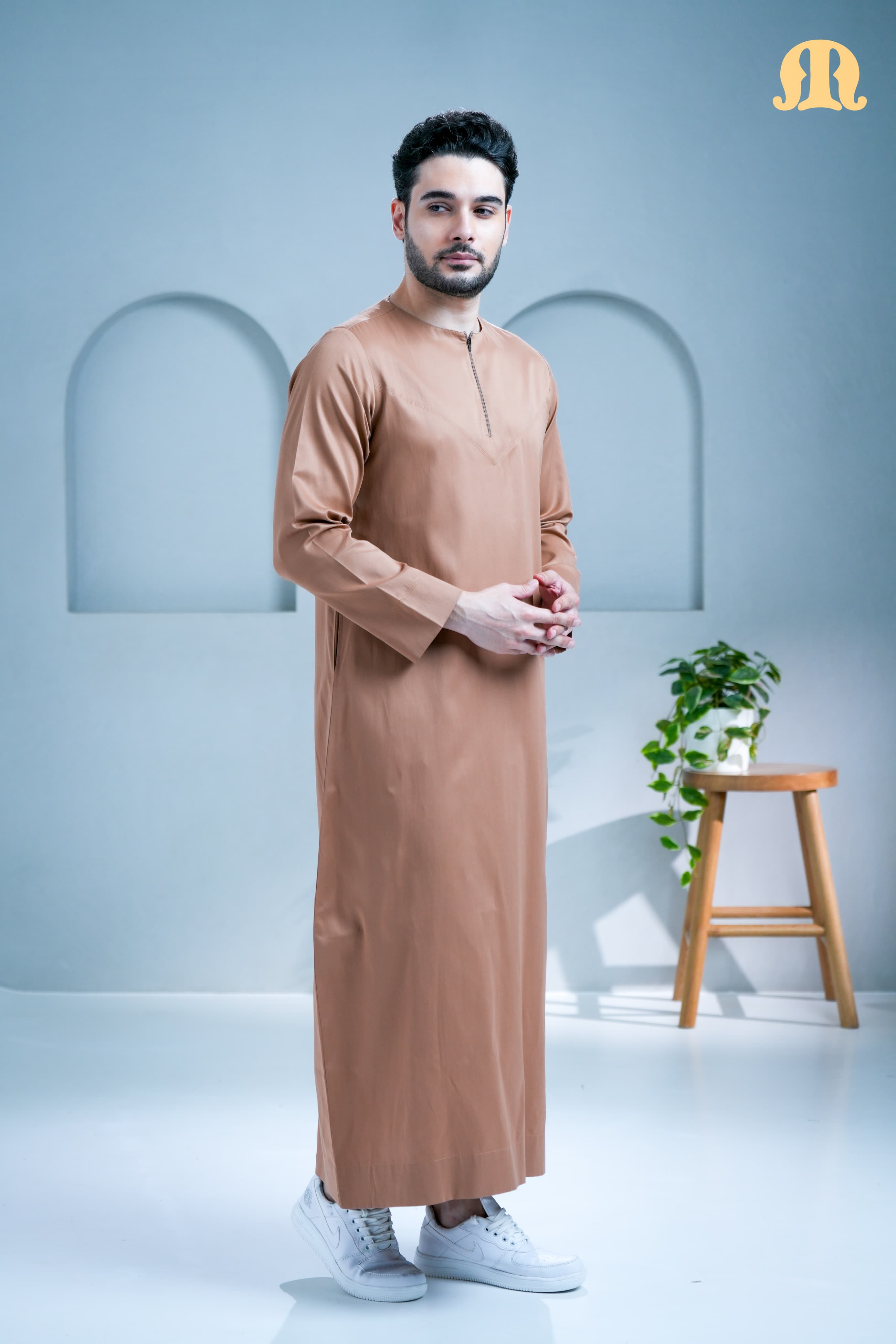 Man wearing a long beige garment standing in a minimalistic room with a plant and stool.